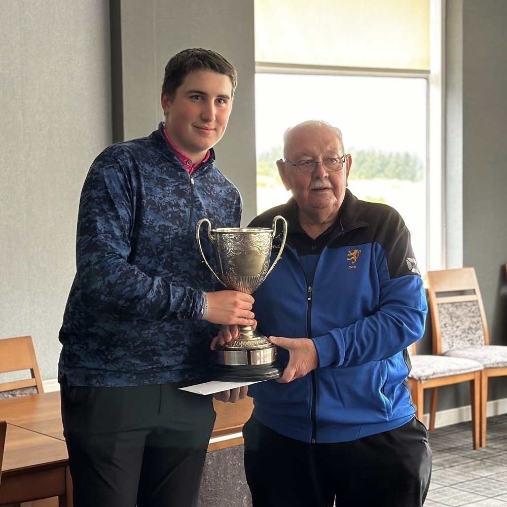 A younger and an older man are standing together indoors, holding a large trophy between them.