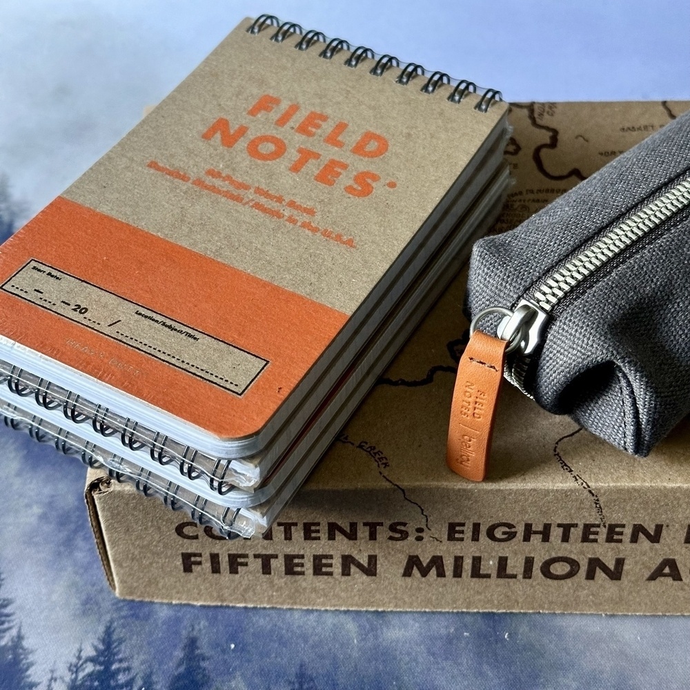 Three spiral-bound Field Notes notebooks and a pencil case rest on top of a cardboard box.