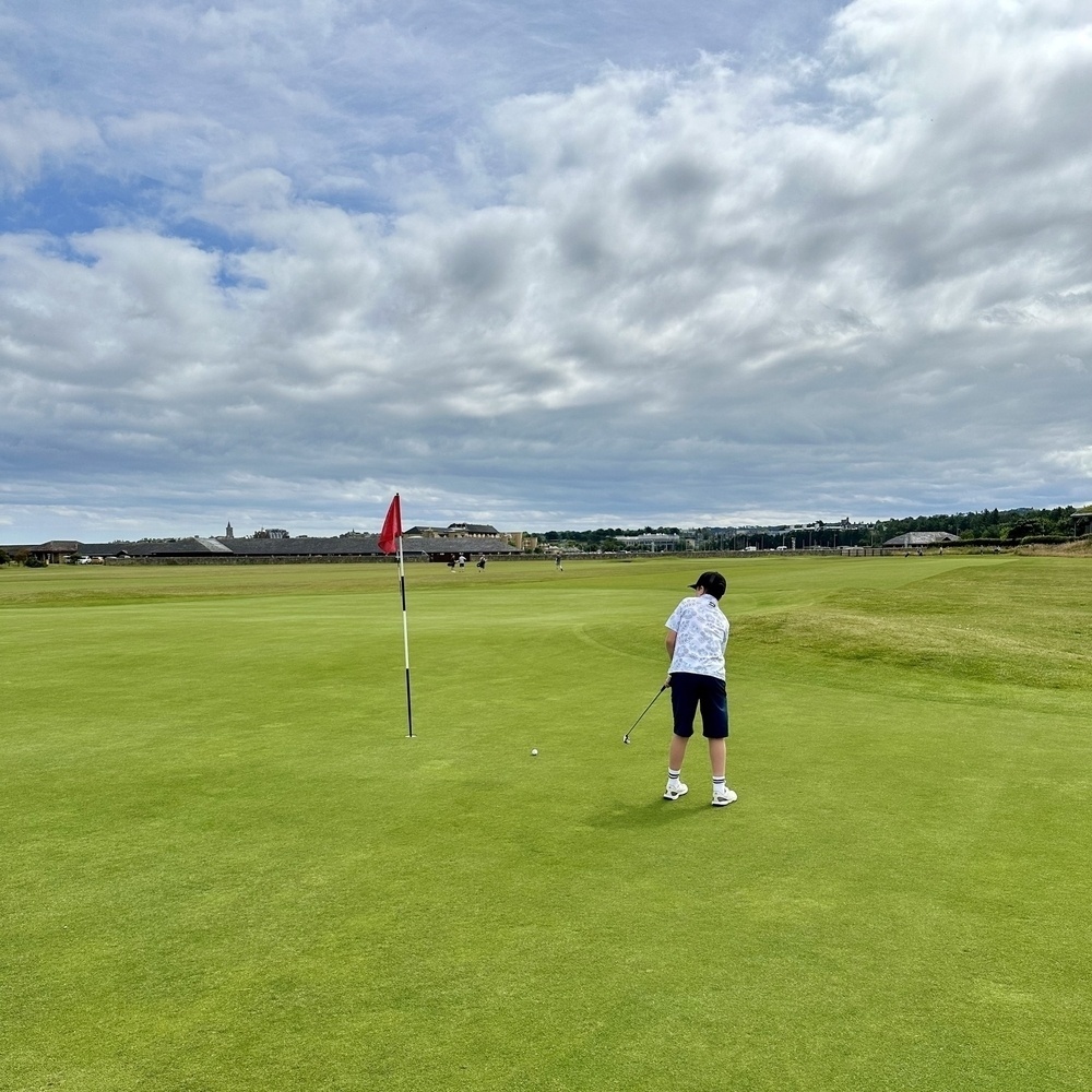 A boy putting out on a golf green with a cloudy sky in the background.