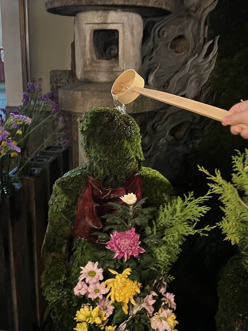 A hand holding a ladle pours water over a moss-covered statue