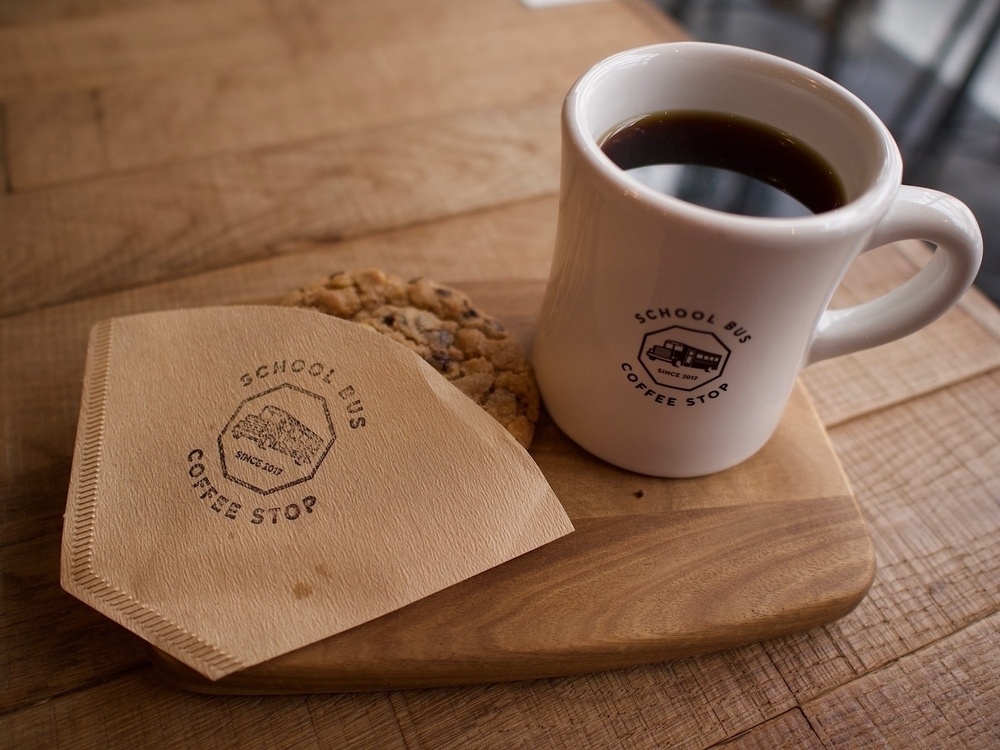 Full coffee cup and a cookie wrapped in a coffee filter. Both the filter and the cup have the School Bus Coffee Shop logo on them