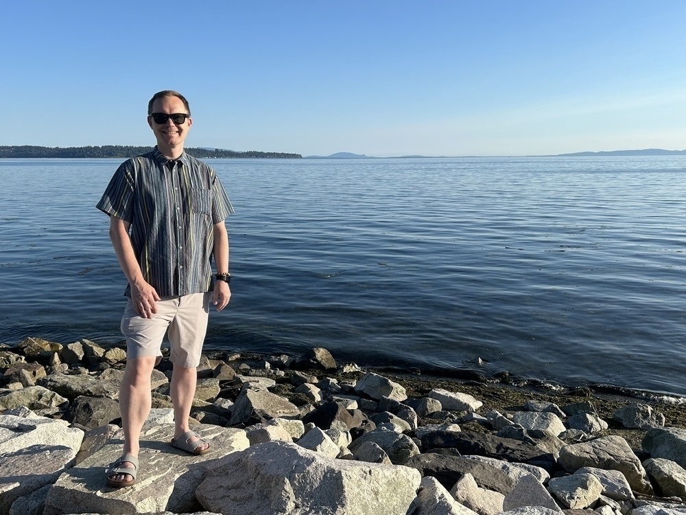 Chad stands on a large rock on the waterfront. In the distance you can see the US shoreline
