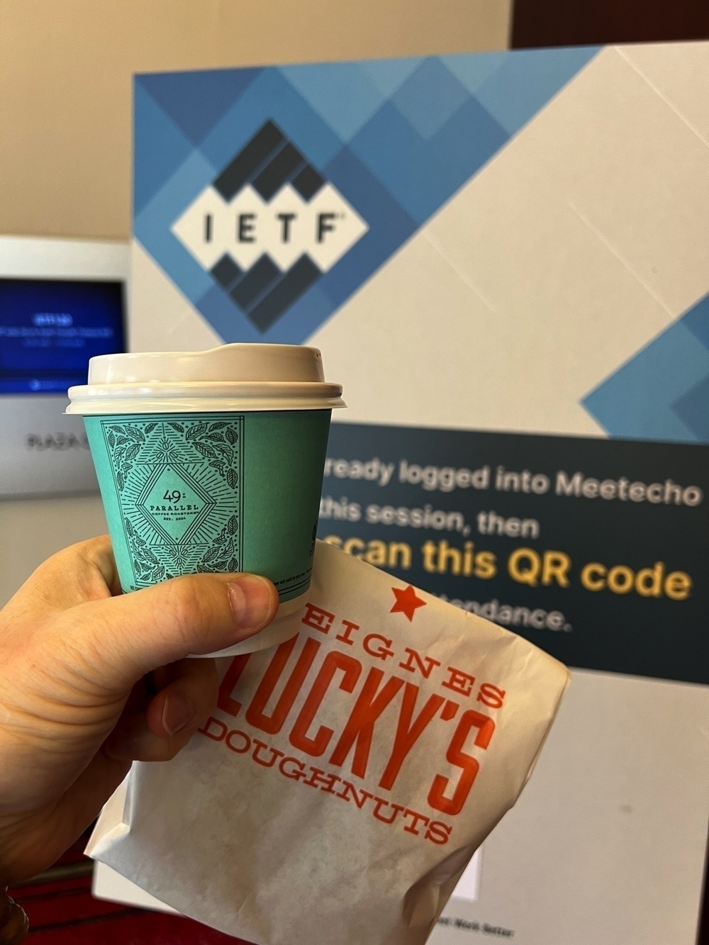 Chad holds a Parallel 49 coffee and bag of Lucky’s donuts in front of an IETF meeting sign