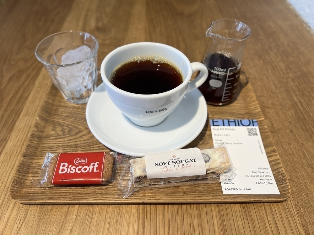 A coffee cup with the words “Life is SÖT”. A Mario beaker has 90mL more of coffee to the right. To the left is a cup with a large ice cube for making ice coffee. A paper label describes the roast as Ethiopian. On the tray is a package of nougat and some Biscoff cookies