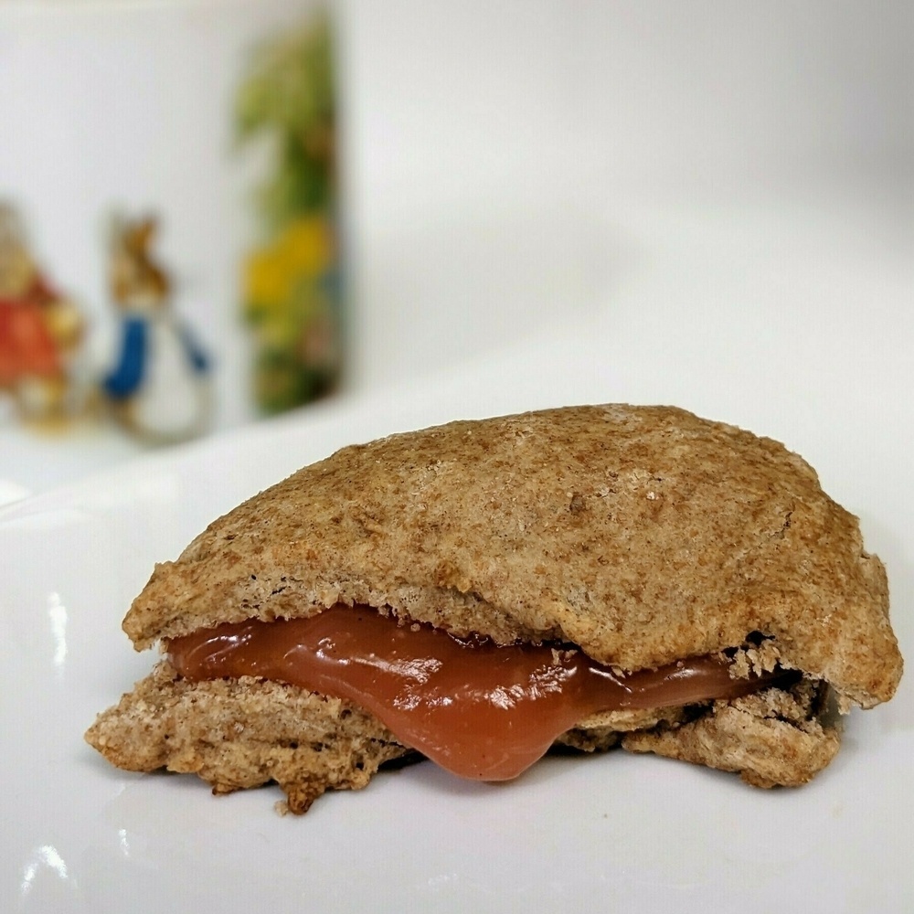 A close up of a brown triangle shaped scone sitting on a white plate. The scone is sliced in half and filled with a dark pink curd that is dripping out the sides. The plate is on a white counter with a white wall behind it. There is a Peter Rabbit mug of tea in the background.