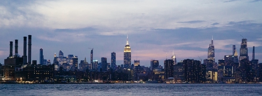 A sunset over Manhattan as seen from Williamsburg. The sky is awash with pastels. The city is full of lights on otherwise dark buildings.