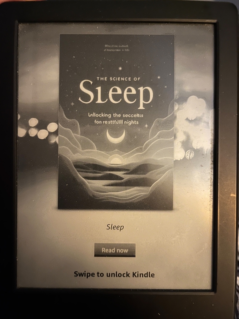 A Kindle screen displaying the cover of a book titled “The Science of Sleep.” The cover features a stylized landscape with mountains, a crescent moon, and stars under the title. Below the image, a button labeled “Read now” is present. There’s a subtitle the looks vaguely like “ullocking the secceltes fon resttfulll nights”