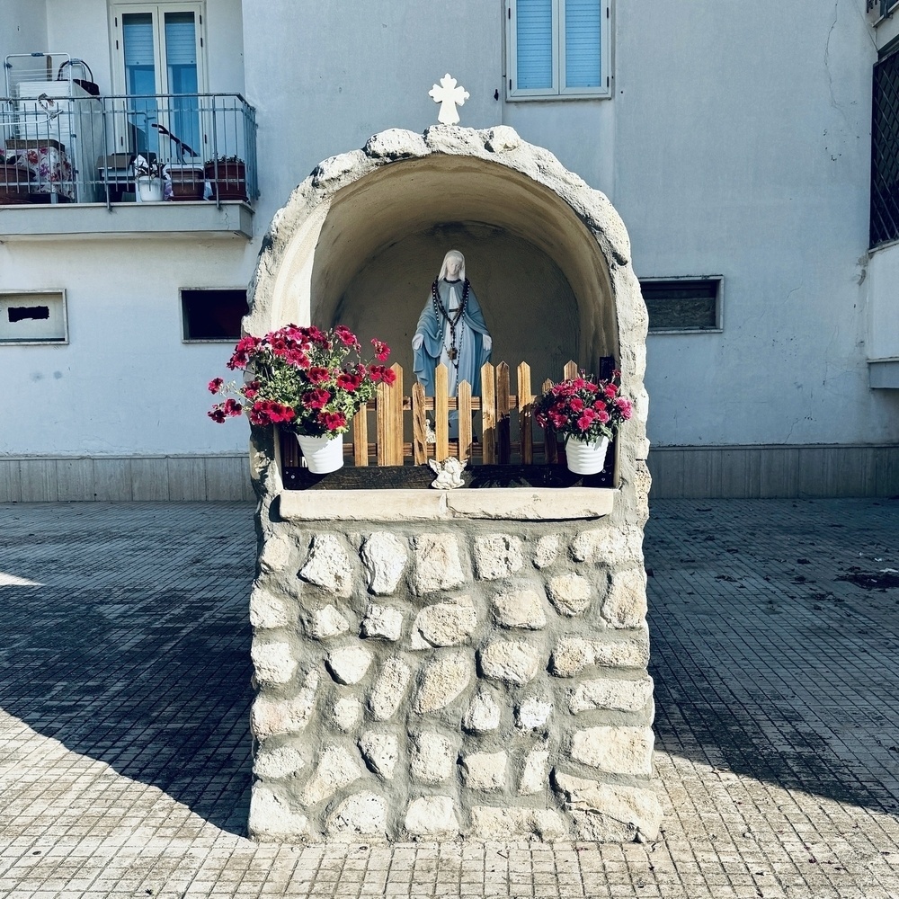 Shrine to Mary in a street. It's made of bricks and there is a brown fence in front of Mary.