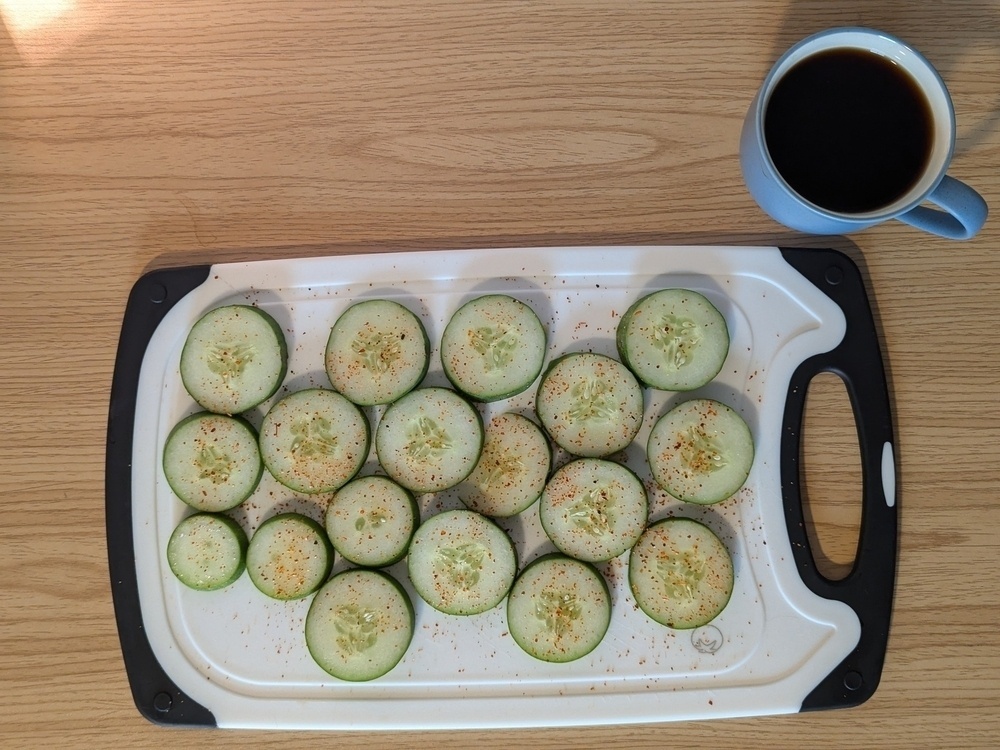 circular slices of cucumber on a cutting board sprinkled with a red seasoning. a cup of coffee sits in the upper right of the frame.