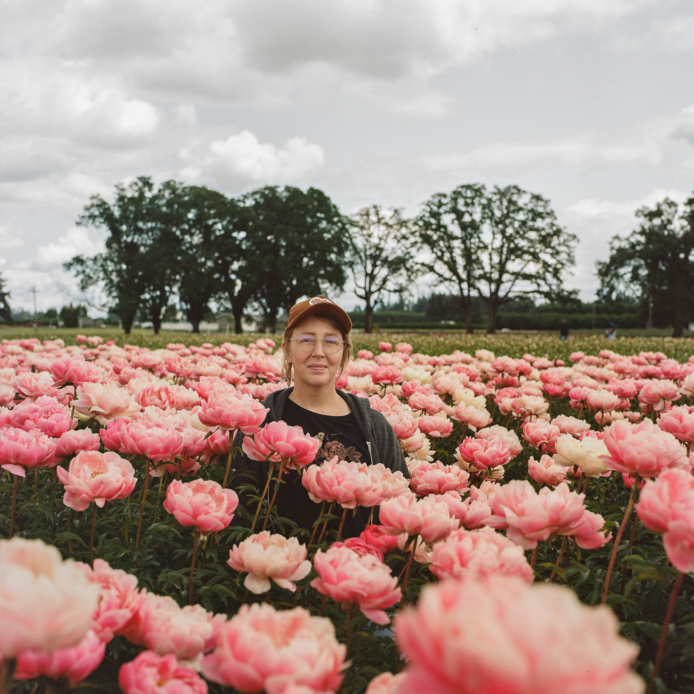A woman wearing a baseball cap stands in the middle of a blooming field filled with pink peonies. There are trees in the background and a cloudy sky overhead.