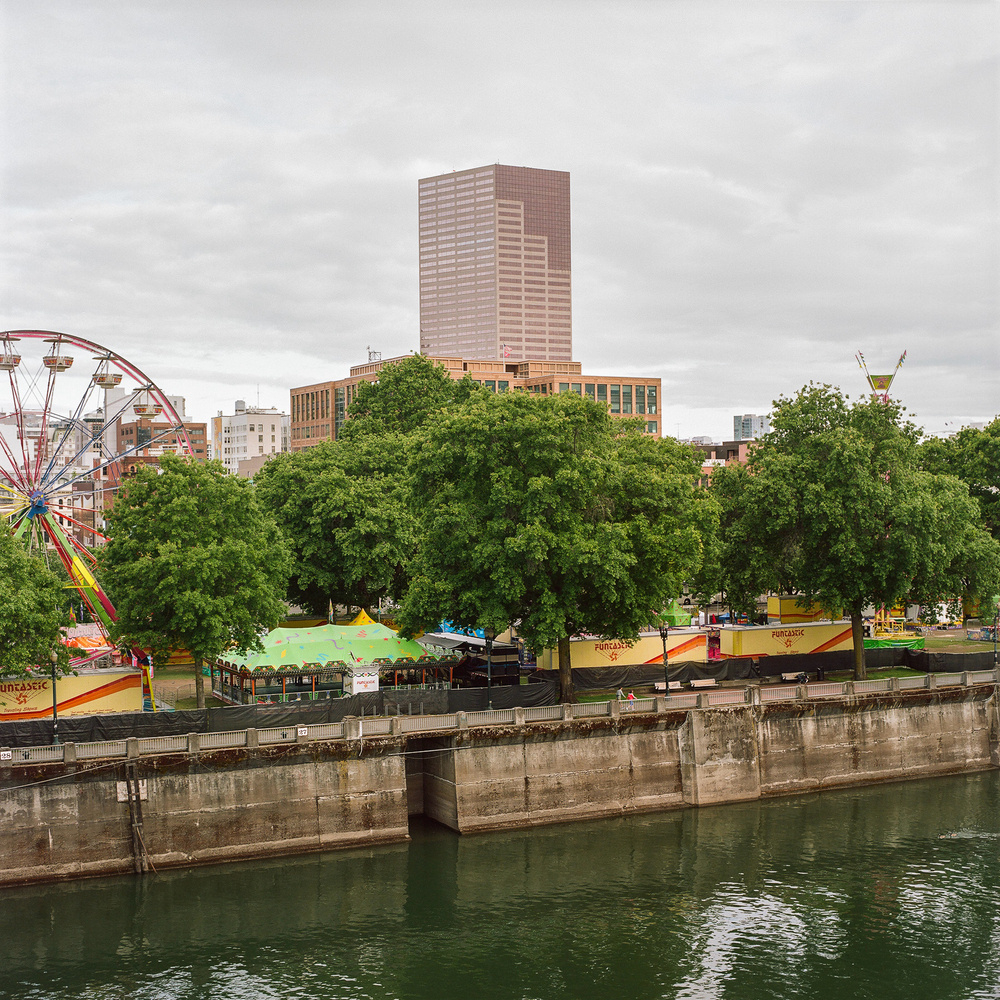 An urban riverside scene with a carnival in progress. The image showcases a Ferris wheel, various colorful tents, and carnival rides, partially obscured by large trees. A calm river runs in the foreground, with a concrete embankment, and a tall building in the background. The sky is mostly cloudy.