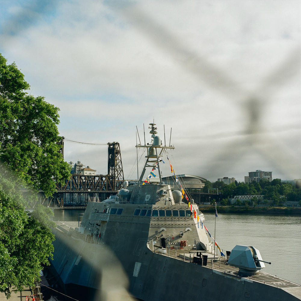 A modern naval ship docked by a river with a steel bridge and an urban skyline in the background. The ship is adorned with signal flags. Green trees are visible on the left side, and part of the image is blurred by an out-of-focus fence.