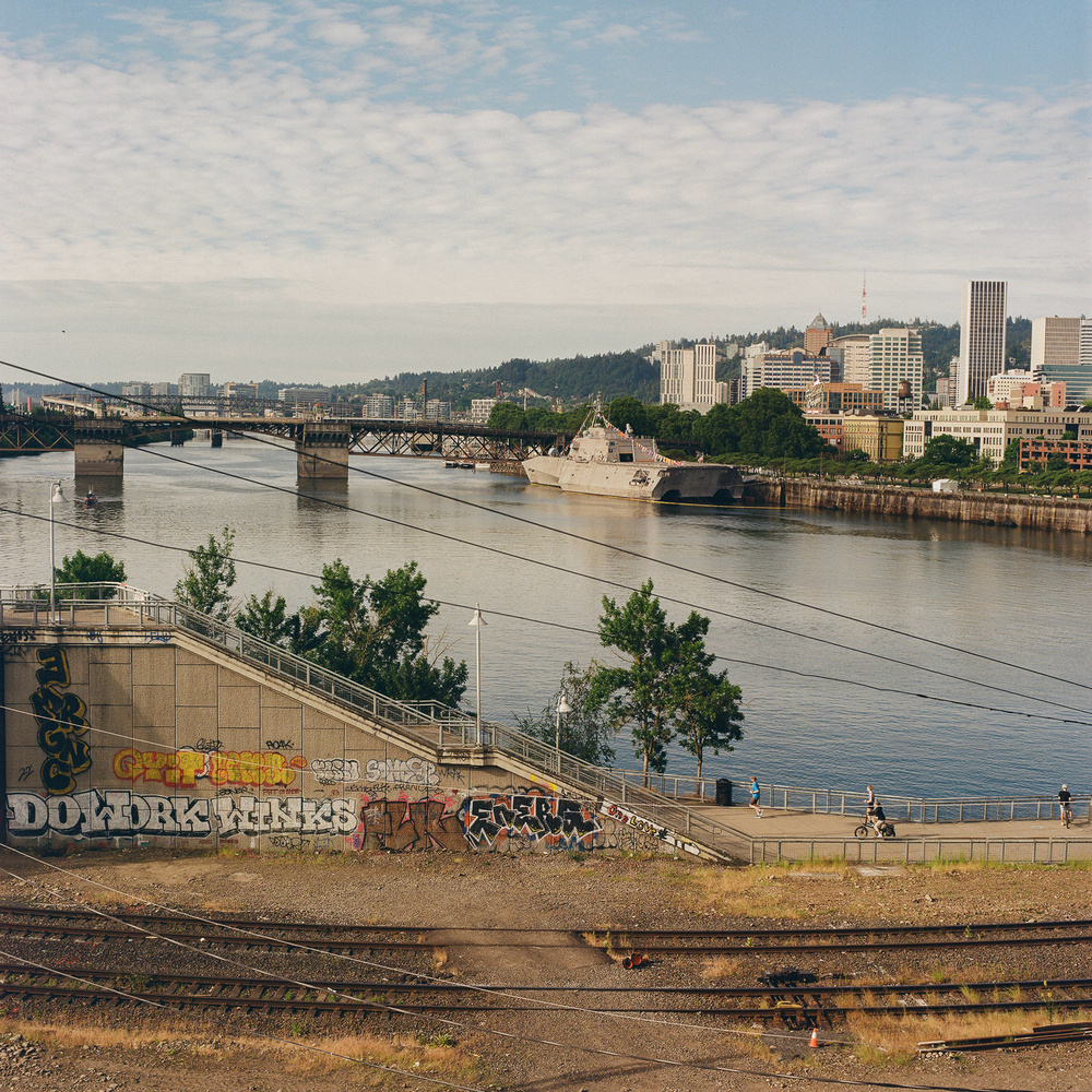 A river with a naval ship docked on the right side. The background features a bridge and an urban skyline with various buildings. The foreground shows railway tracks, graffiti-covered walls, and a paved pathway with people.