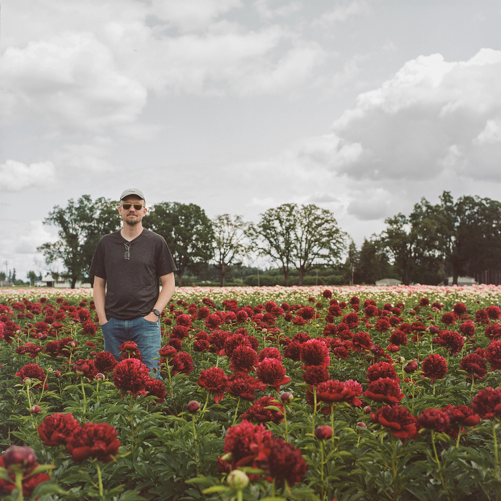 A man stands in a field of vibrant red peonies. The sky above is cloudy, and there are trees in the background.
