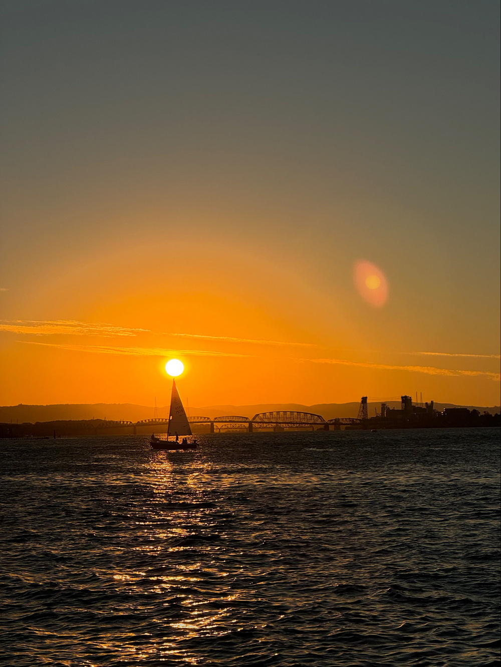 A sailboat on a body of water during sunset with the sun near the horizon. The sky is a mix of orange and yellow hues, with the sunlight reflecting off the water. There is a bridge and industrial structures visible in the background.