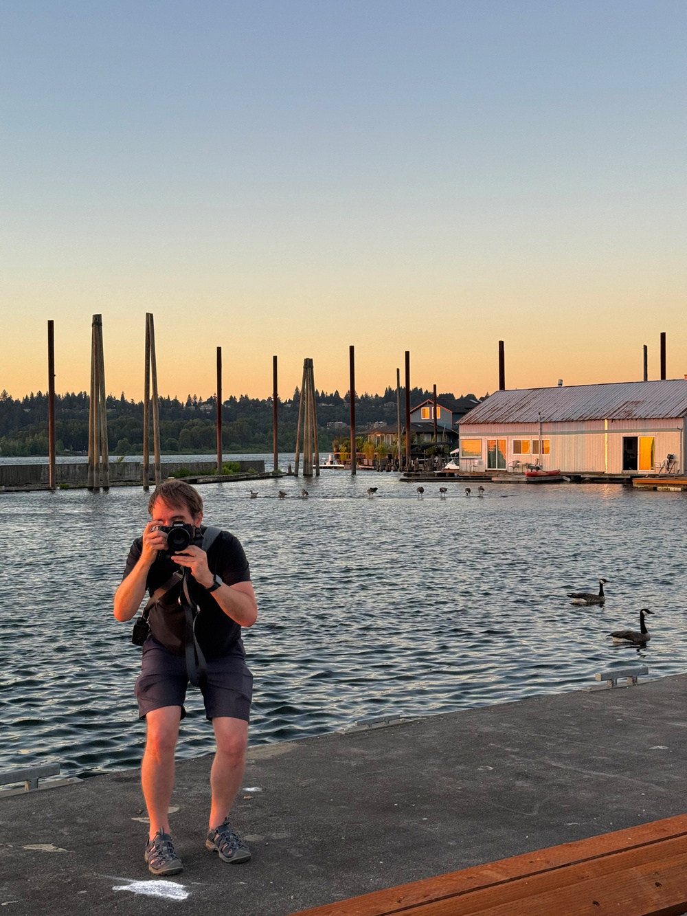 A person is crouched near a waterfront, taking a photo of the photographer. The background features a calm body of water, several geese, and a house with a tin roof. The sky is clear with a gradient from blue to orange during sunset.