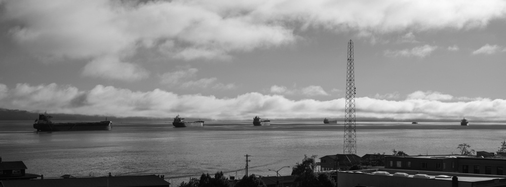 A black and white photo shows several large cargo ships anchored in calm water under a sky filled with clouds. In the foreground, there are rooftops and structures, including a tall metal tower, along a shoreline.