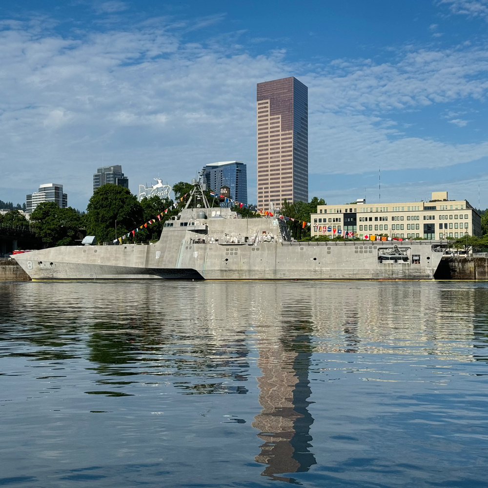 A military ship docked in a city harbor, with decorated flags adorning its deck. Urban buildings and a distinctive tall skyscraper are visible in the background. The water reflects the ship and buildings. The sky is partly cloudy.