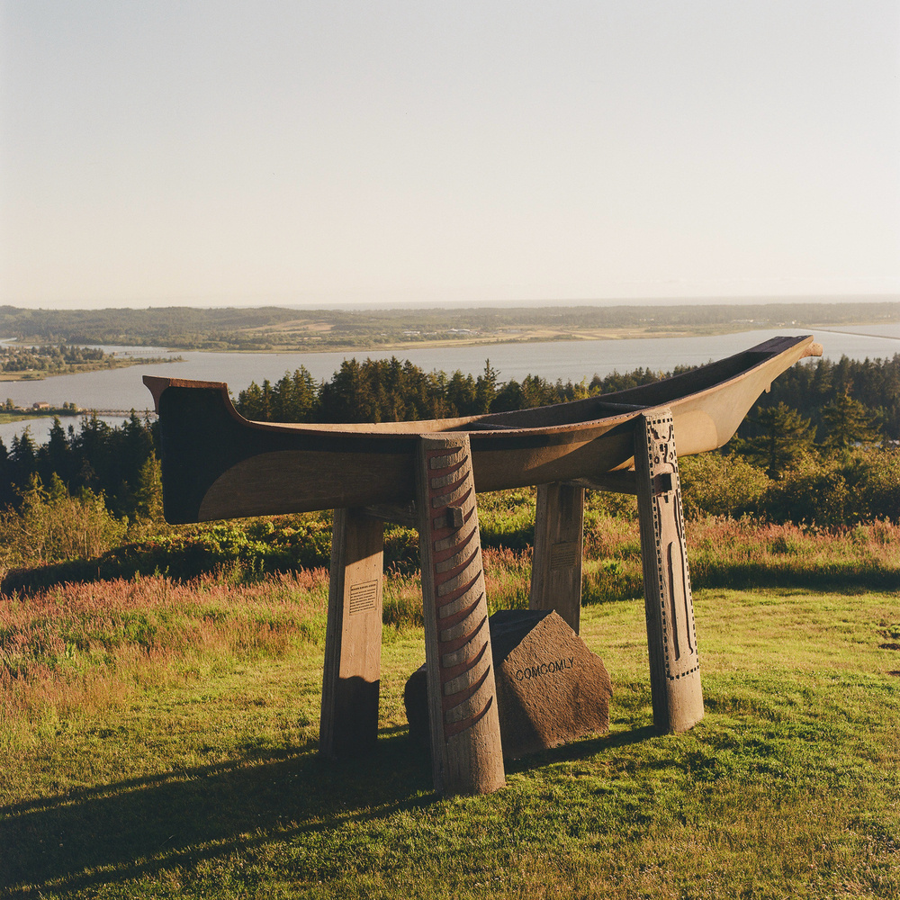 A wooden sculpture featuring a traditional canoe design, supported by four intricately carved vertical posts, is set on a grassy hilltop. It overlooks a scenic landscape with a forested area, a river, and distant terrain under a clear sky. A rock under the canoe has carved text that reads, “Comcomly”.