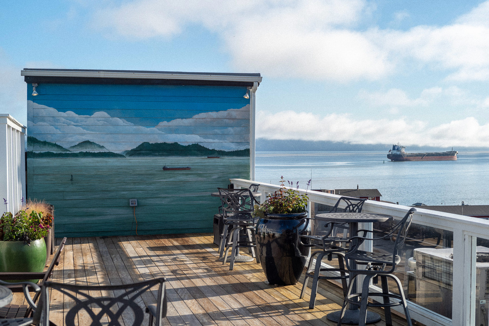 A rooftop deck with wooden flooring, metal chairs, and tables, surrounded by potted plants. The deck overlooks a body of water with a visible cargo ship in the distance. A mural depicting a similar scene is painted on a wall.