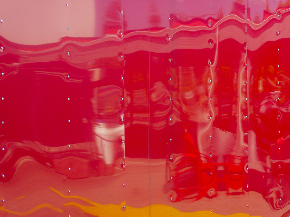 A distorted reflection of a street scene on a shiny, red metallic surface, adorned with rivets, creating a wavy and abstract visual effect.