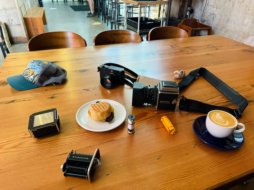 A wooden table with a vintage camera, a hat, a plate with a sandwich, a coffee cup with a latte art, two film camera accessories, an exposed film roll, and a small wrapped film roll, in a cafe setting.