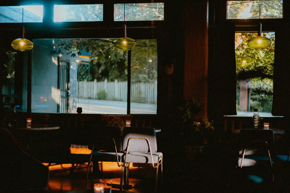 A dimly-lit, cozy interior of a bar with vintage chairs and tables. Soft light filters through large windows, enhancing the warm ambiance. Green hanging lamps illuminate parts of the room. A soft glow from small candles on the tables adds to the intimate feeling.