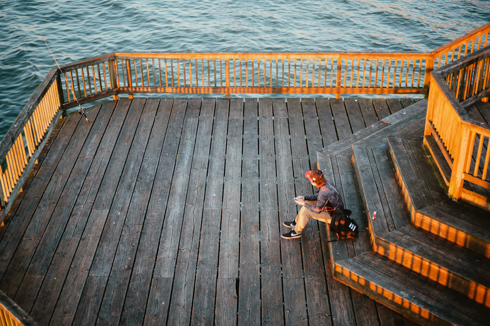 A person sits on wooden steps next to a railing, facing a wooden deck and a body of water. A fishing rod is seen propped against the railing. The scene is lit by warm sunlight.