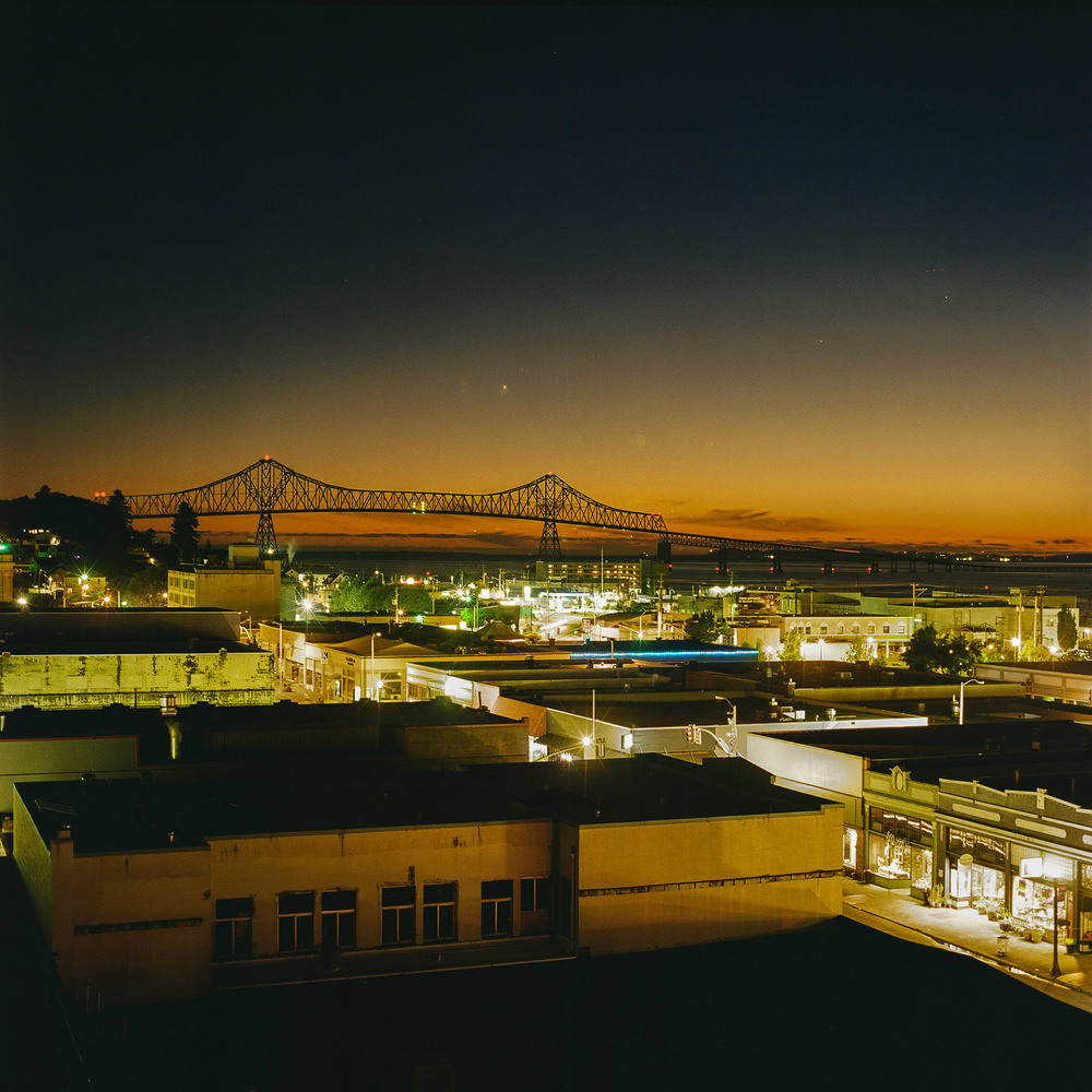 Nighttime cityscape of Astoria, Oregon with illuminated buildings in the foreground and the prominent Astoria-Megler Bridge spanning the Columbia River in the background, against a twilight sky.
