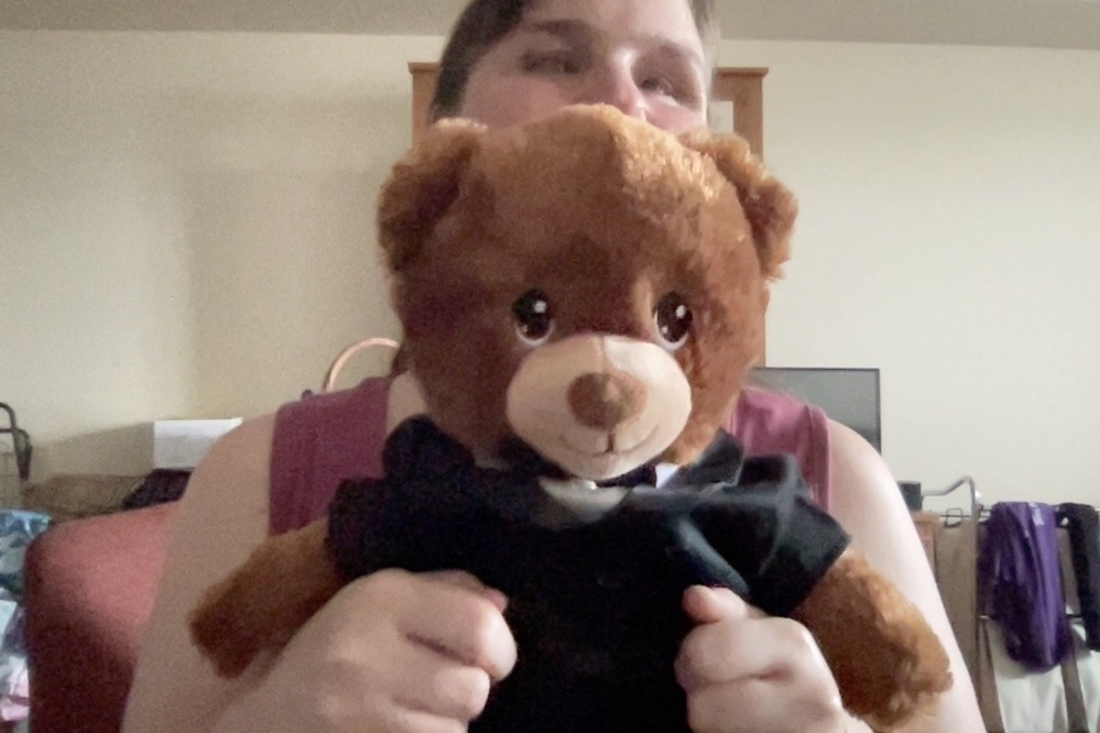 The picture primarily features a cute brown teddy bear wearing a black suit with a bow tie. The teddy bear has a friendly face with black eyes and a little nose. The background of the picture shows a room with a plain wall, and there is some furniture and clothes hanging visible in the background.