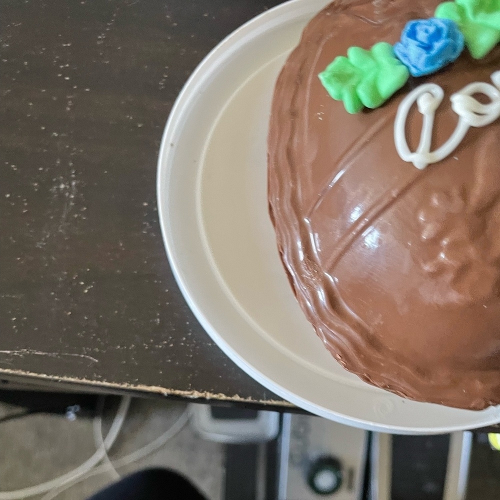 The image features a delicious chocolate easter egg with white frosting, sitting on a white plate. The egg is beautifully decorated with green and brown accents, giving it a festive appearance. The plate with the egg is placed on my desk. You can also see several other items in the photo, including a coffee mug and my apple TV remote.