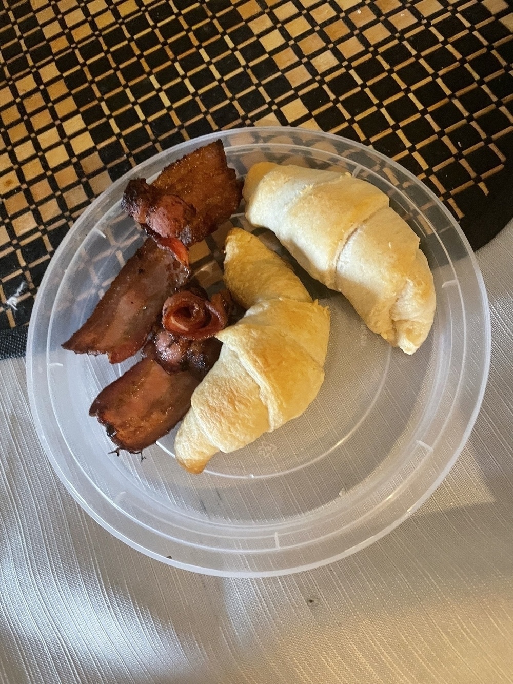 A transparent plate sitting on a checkered tablecloth. On the plate, there are 2 freshly-baked croissants.  They are flaky, soft, and golden brown. There are also several pieces of crispy-looking bacon on the plate with the croissants. ￼￼