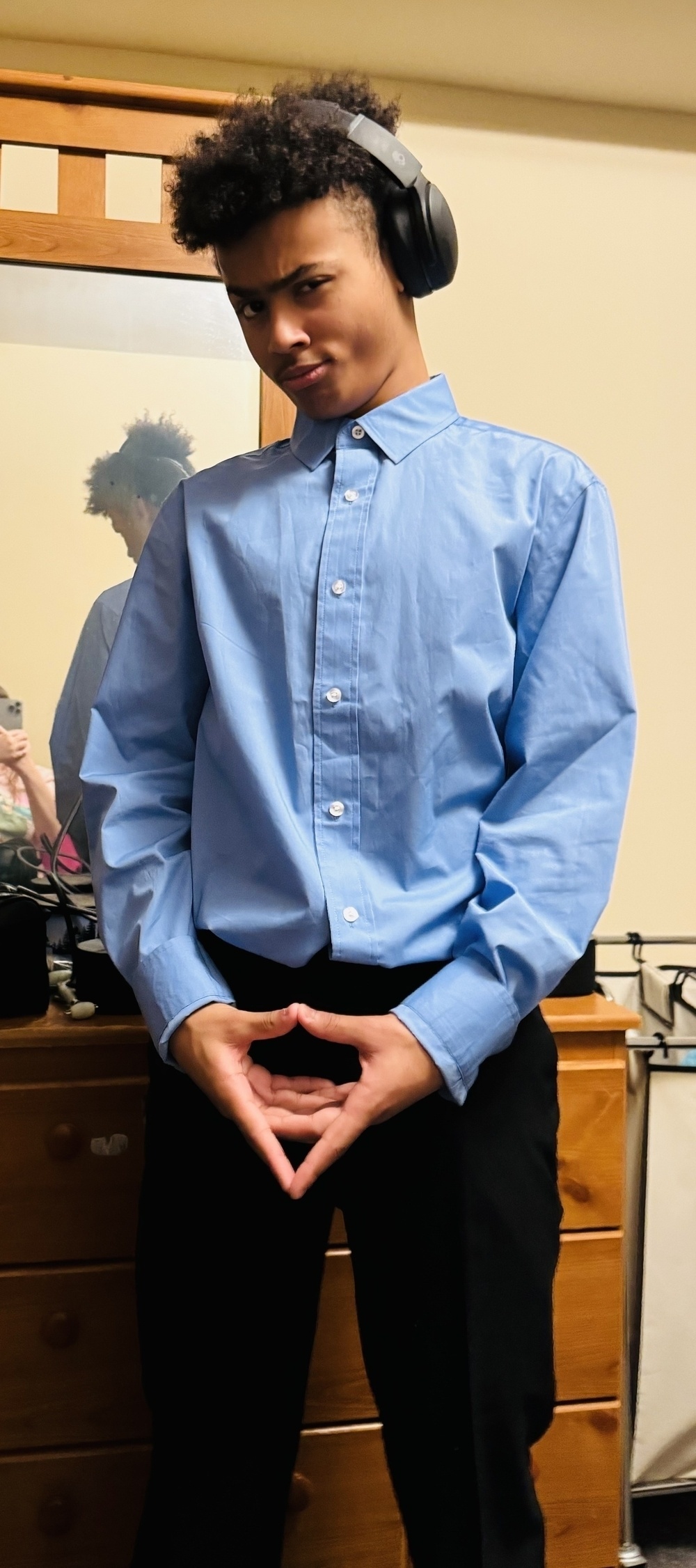 The picture shows a young man standing in a room. He has curly hair and is wearing large headphones over his ears. He is dressed in a light blue button-up shirt and black pants. He is making a heart shape with his hands by joining his thumbs and fingers. Behind him, there is a wooden bed frame, a mirror reflecting his back, and a wooden dresser with drawers. There are also some items on the dresser, and a part of another person is visible in the background. The young man is looking at the camera with a slight smirk on his face.