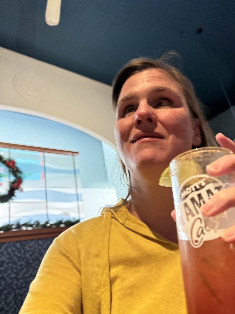 I amholding a glass with a reddish drink, a Caesar, one of my favourite cocktails, with a lime slice on the rim. I am smiling slightly and wearing a yellow top. The background is blurred but appears to be an indoor setting with a blue ceiling and some festive decorations visible. The glass has the text "Mott's Clamato Caesar" on it.