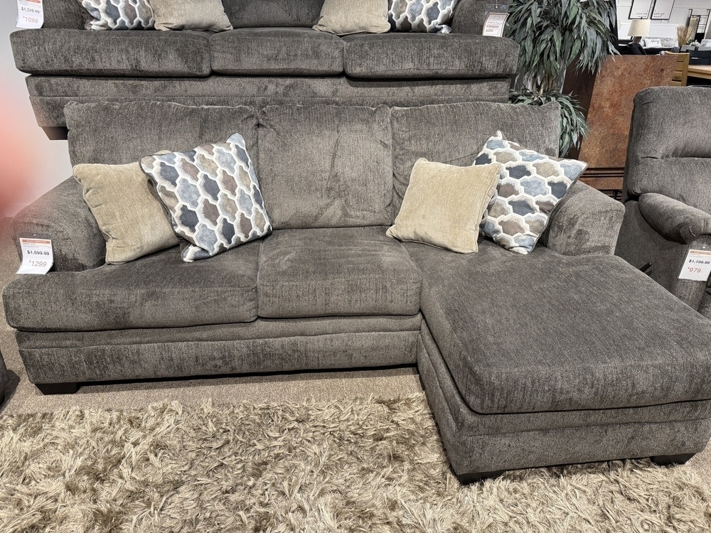 The image shows a gray sectional sofa with a chaise lounge on the right side. The sofa is upholstered in a textured fabric and is accompanied by four decorative pillows. Two of the pillows have a geometric pattern in shades of blue, gray, and beige, while the other two are solid beige. The sofa is displayed in a showroom setting, with price tags visible on the armrests. In front of the sofa, there is a shaggy beige rug on the floor. In the background, there is another sofa stacked on top and a potted plant to the right.