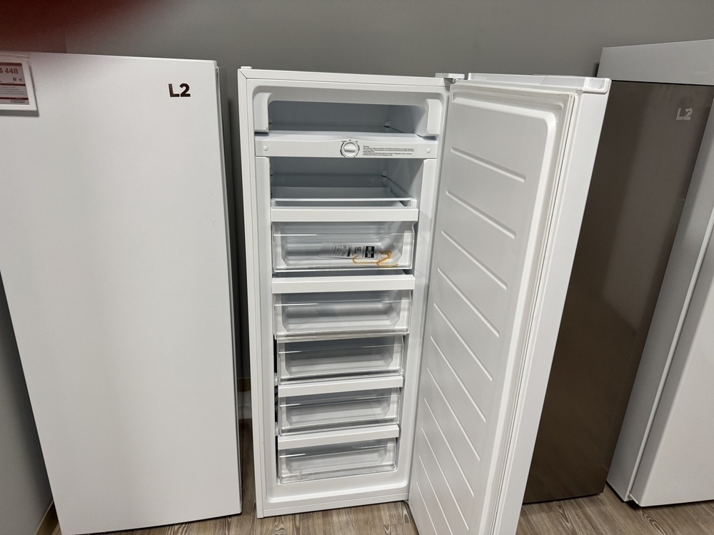 The image shows an open upright freezer with several transparent drawers inside. The freezer door is open, revealing five drawers stacked vertically. The interior is white, and there is a control dial visible at the top. The freezer is placed on a wooden floor, and there are other appliances visible on either side, with one labeled "L2" on the left.