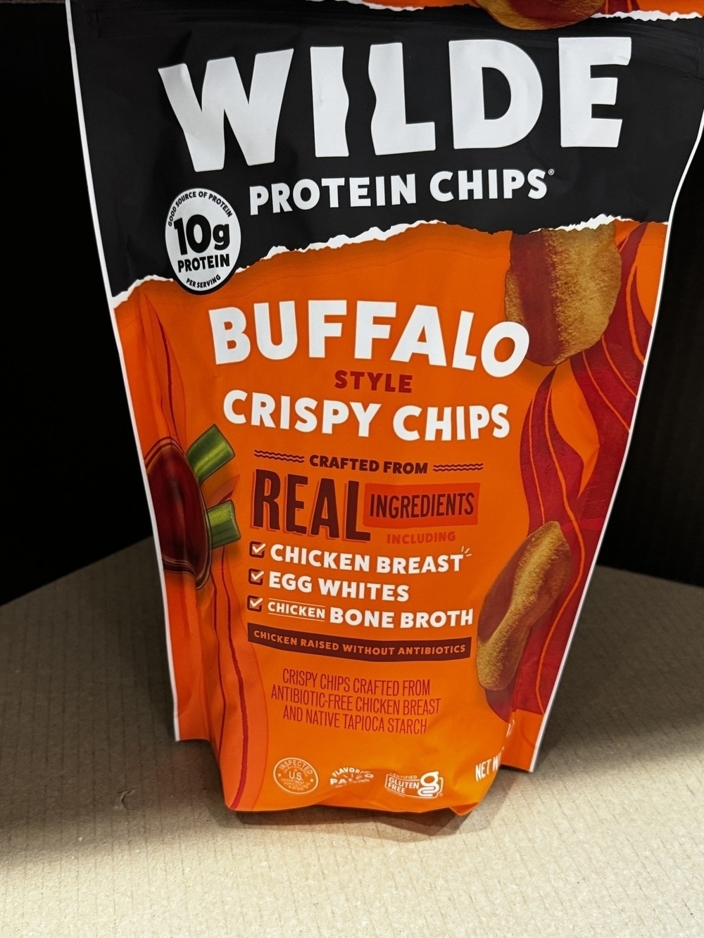 A bag of “Wilde Protein Chips” sits on a cardboard surface, displaying buffalo-style crispy chips made from chicken breast, egg whites, and bone broth, boasting 10g of protein per serving.