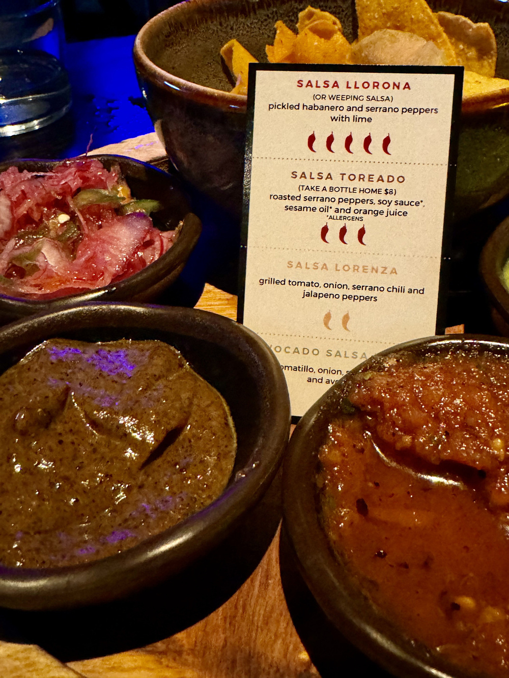 Various salsas in bowls are displayed with a menu detailing their ingredients; tortilla chips accompany them in a dimly lit setting.
Text:
SALSA LLORONA
(OR WEEPING SALSA)
pickled habanero and serrano peppers
with lime
SALSA TOREADO
(TAKE A BOTTLE HOME $8)
roasted serrano peppers, soy sauce*, sesame oil* and orange juice
*ALLERGENS
SALSA LORENZA
grilled tomato, onion, serrano chili and jalapeno peppers
AVOCADO SALSA
tomatillo, onion, cilantro, chili and avocado