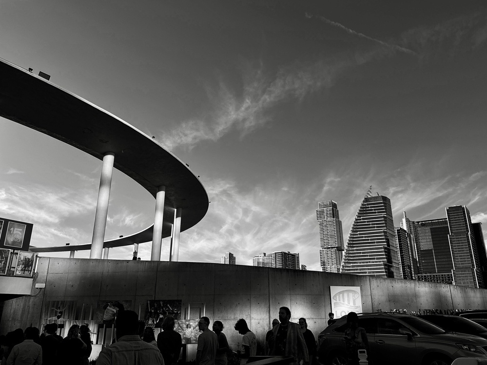 Silhouetted people gather under a curved concrete structure; towering skyscrapers rise in the background against a cloudy sky. The mood is urban and busy.