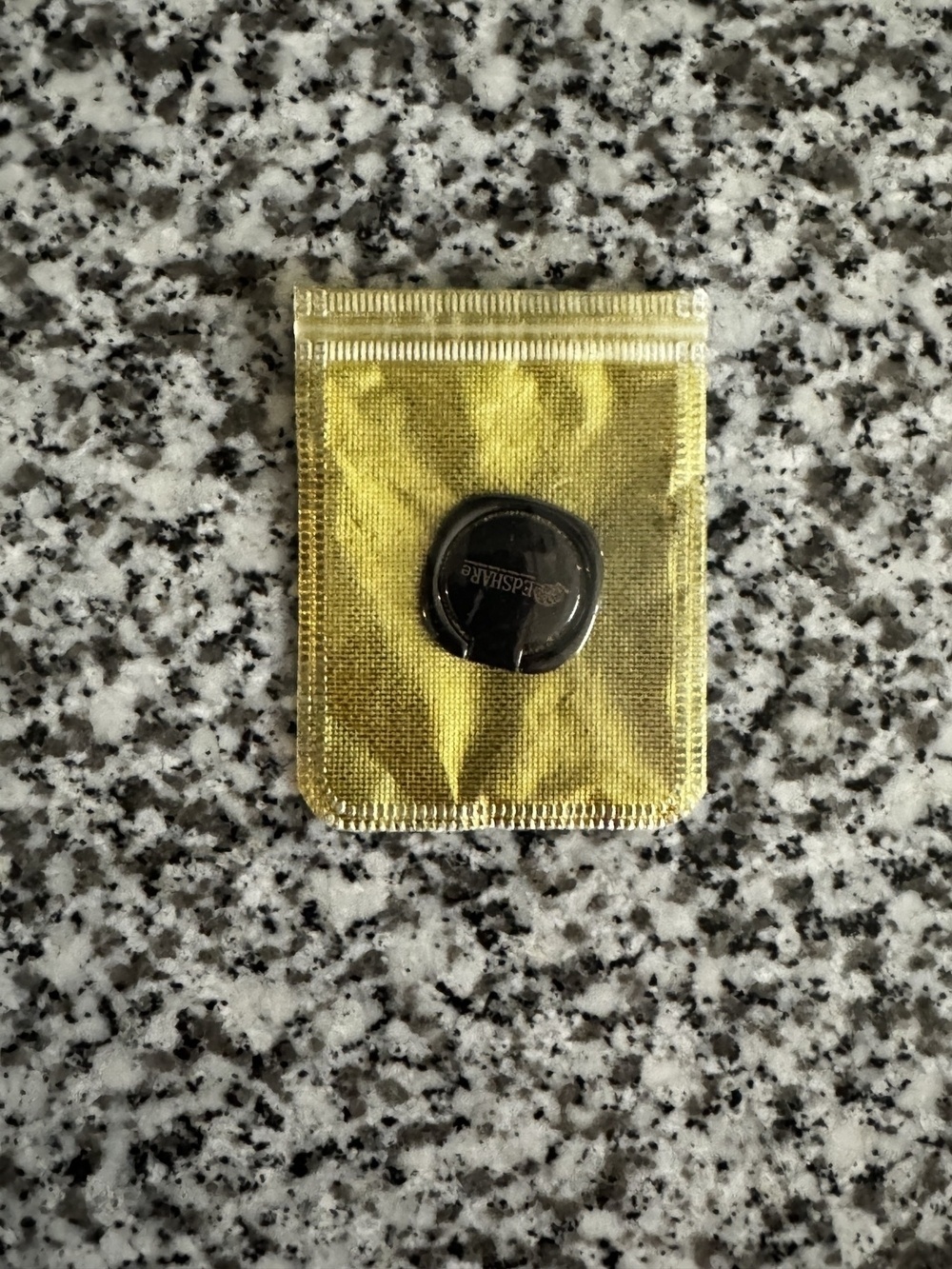 A sealed gold-colored wrapper rests on a speckled counter, containing a circular object embossed with text.