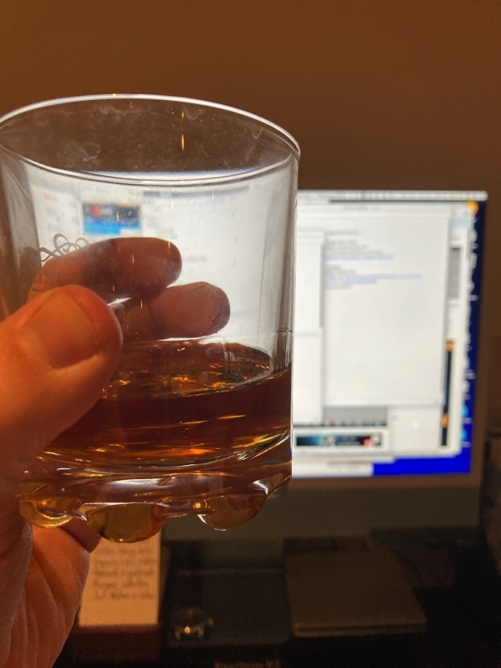 A glass of whisky, held in front of an iMac with the screen blurred in the background.
