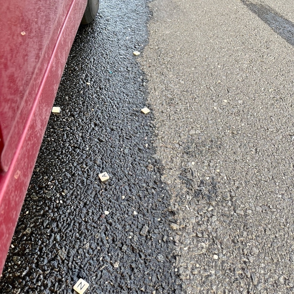 Photo of Scrabble game tiles scattered on a road next to a red car.