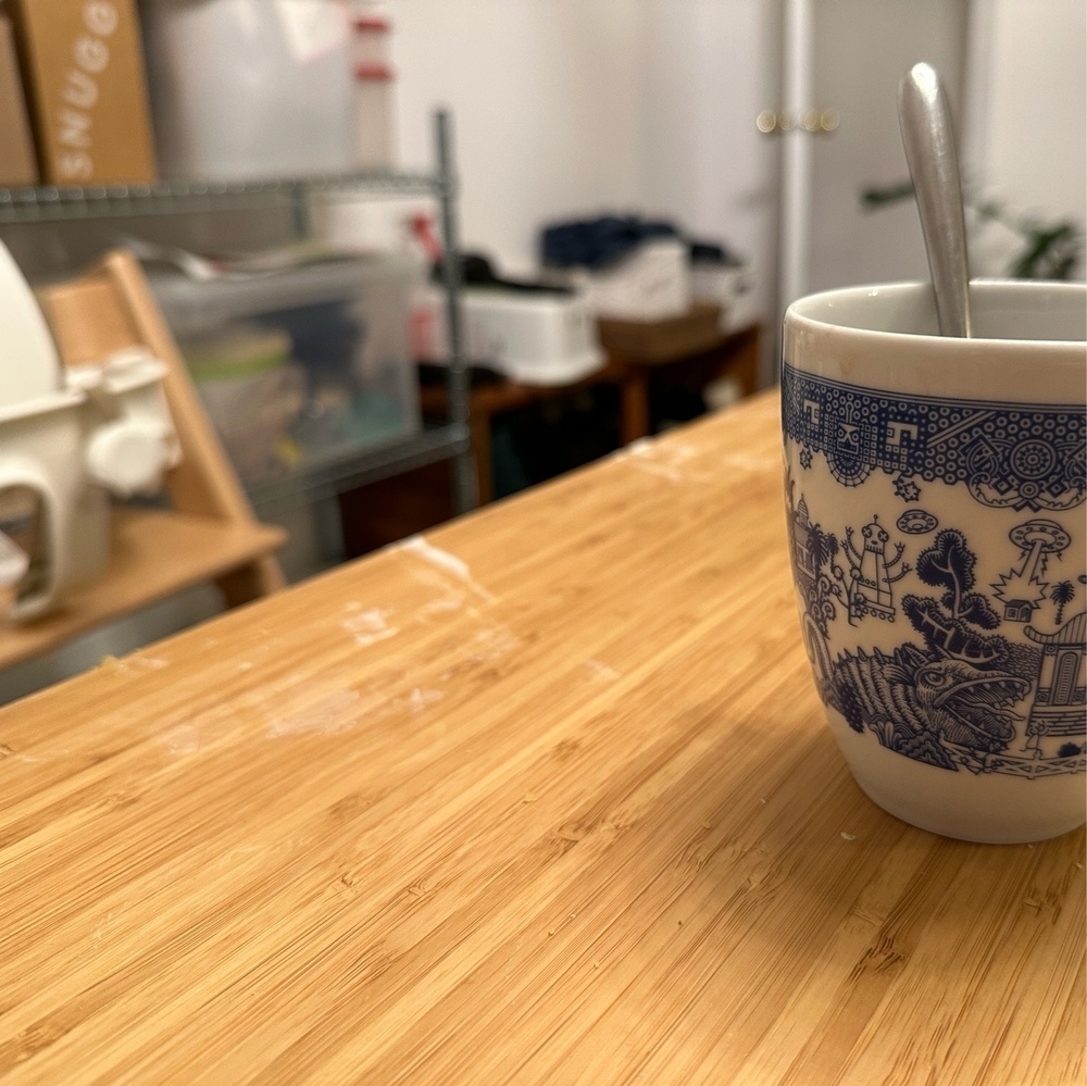 Photograph of bamboo-top dining table, mug with spoon inside of it, and a mess of yogurt on the edge of the table.