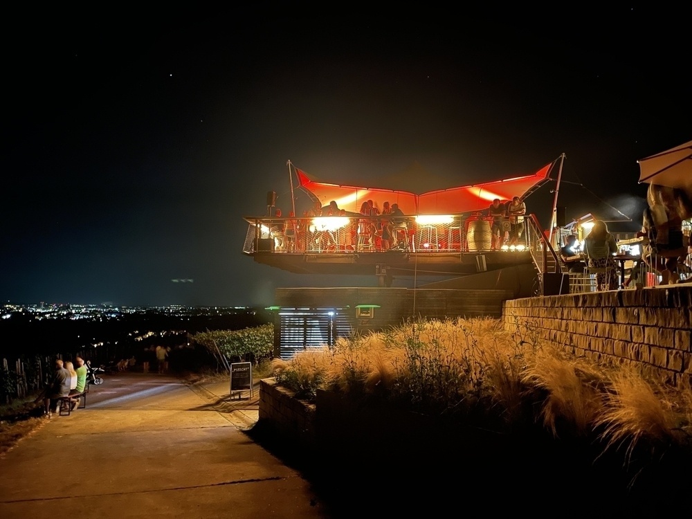 A group of people gathers at a brightly lit, elevated outdoor venue with a red canopy at night, overlooking a cityscape.