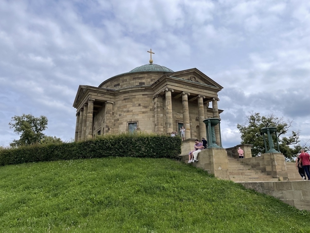 A neoclassical-style building with a domed roof and a cross on top is situated on a grassy hill with people standing near it.