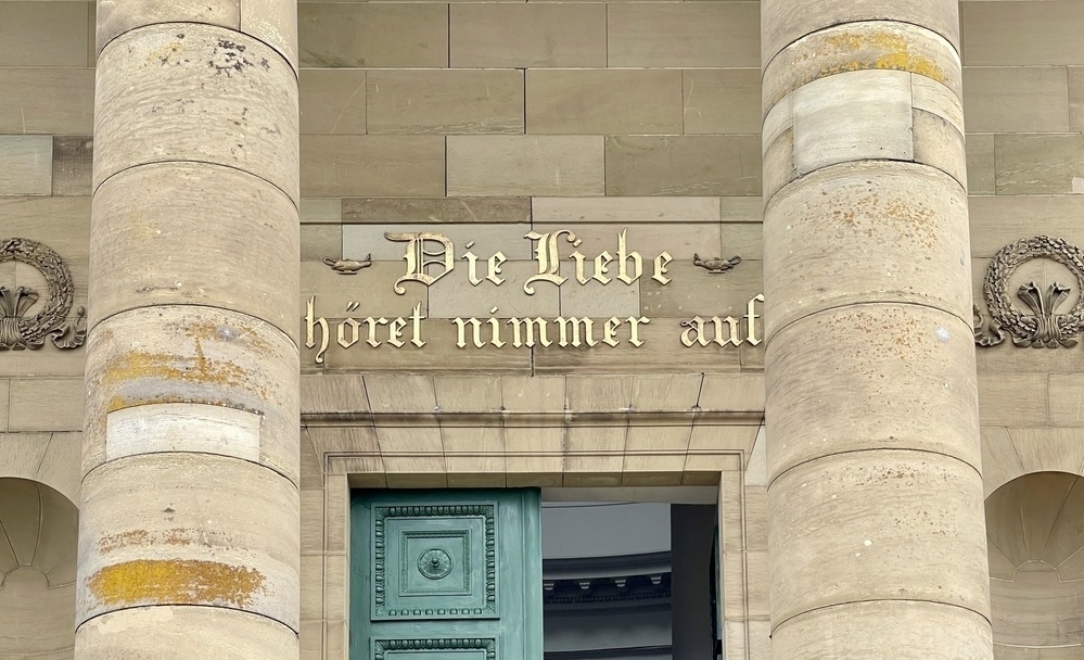 A close-up view of a historic building’s entrance features grand stone columns, intricate detailing around a teal door, and an inscription in German above the doorway: „Die Liebe höret nimmer auf“ - „The love never ends“