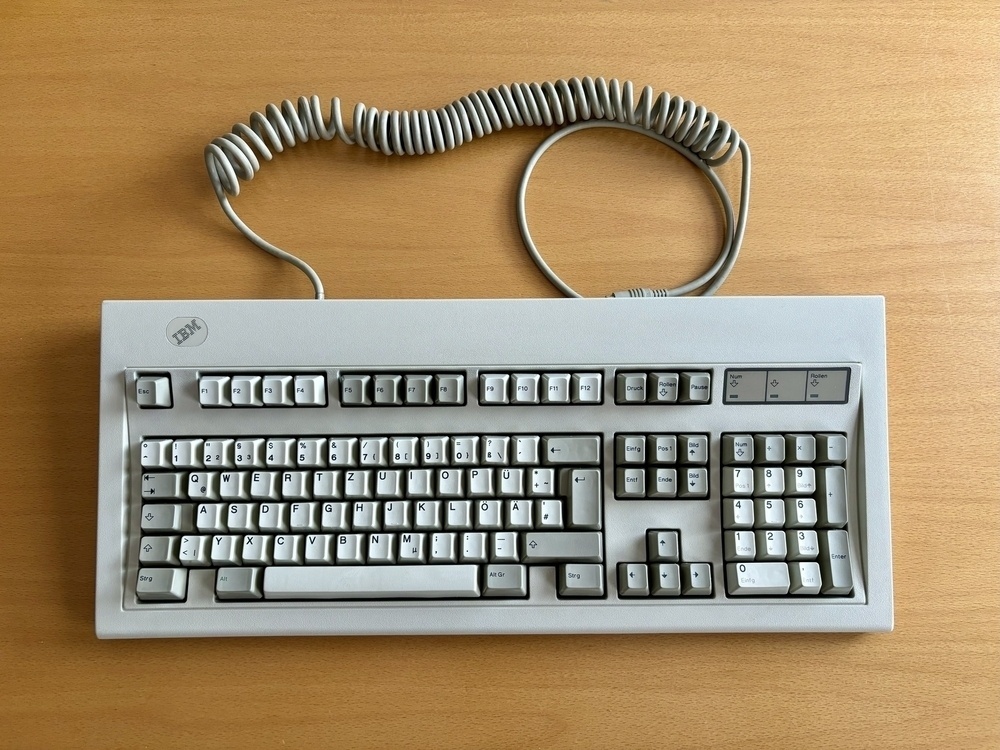 A vintage IBM Model M mechanical keyboard from 1988 with a coiled cable is placed on a wooden surface.