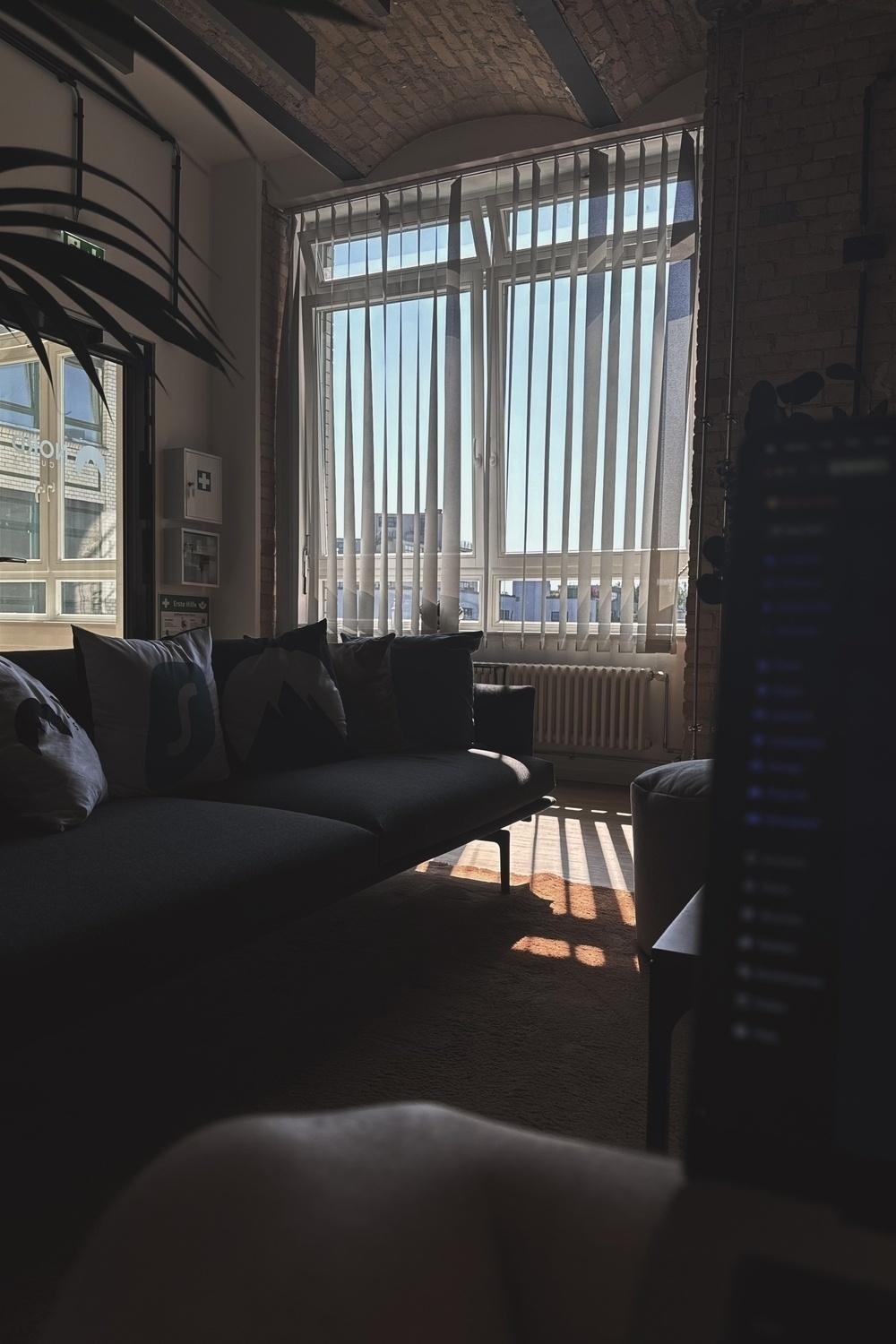 A cozy, sunlit office room features a dark couch with pillows in front of a large window with vertical blinds partially open.