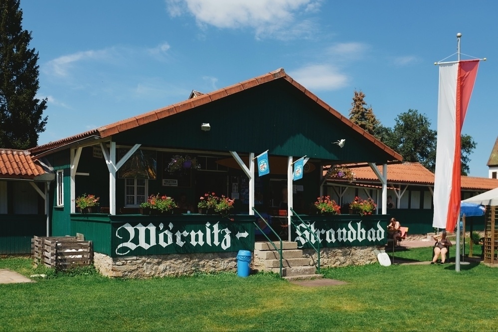 Green and white wood kiosk at the Wörnitzstrandbad river beach. The kiosk is decorated with red flowers and there’s a tall red and white flag hanging from a mast and a couple of people sunbathing.