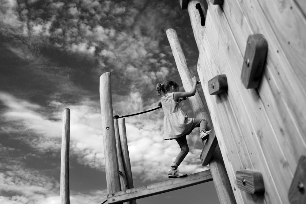 Black-and-white photo of a child climbing on a wooden playground jungle gym against a background of a clouded sky.